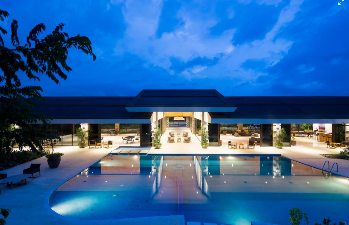A nighttime view of a hotel pool area with a large pool, outdoor furniture, and a building in the background.