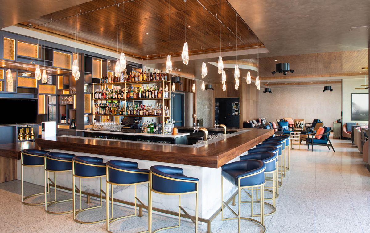 A well-lit bar with a wooden counter, blue stools, and gold accents.